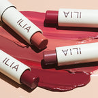 ILIA Balmy Tint Hydrating Lip Balm – cremefarbene Tube mit farbigen Stiften, rosafarbener Hintergrund – Lippenpflege