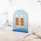 Spectral Orange Ohrringe – orangefarbene geometrische Formen auf blauem Acrylhalter, Marmorhintergrund – Schmuck