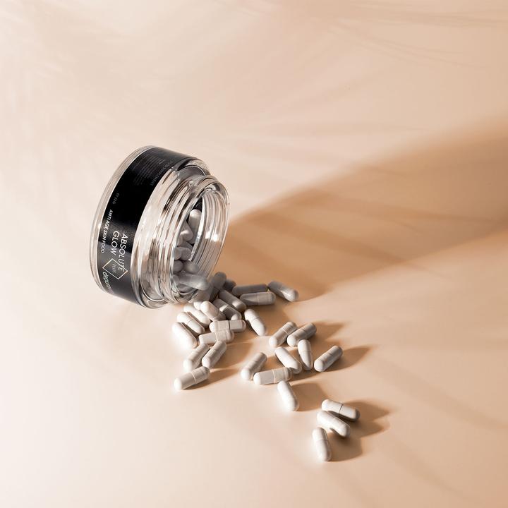 Absolute Glow Plus – durchsichtige Glasdose mit schwarzem Etikett, weiße Kapseln, beige Hintergrund – Anti-Aging