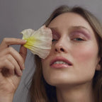 Cream Blush Blossoming – Model mit rosafarbener Wange, Blütenblatt vor Auge, grauer Hintergrund – Gesichtsmake-up