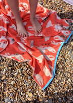 Lobster Bio-Baumwoll-Strandtuch – leuchtend orange mit weißen Hummermotiven, blauer Saum, auf Kieselstrand – Strandaccessoires