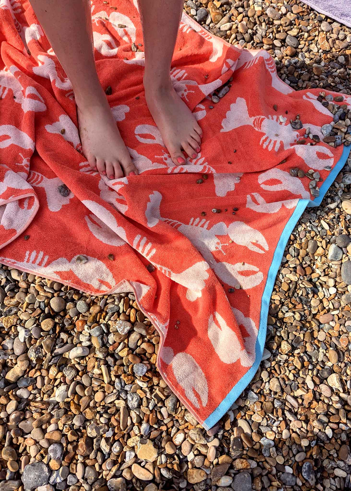 Lobster Organic Cotton Beach Towel – vibrant orange with white lobster print, blue border, on pebble beach – beach accessories
