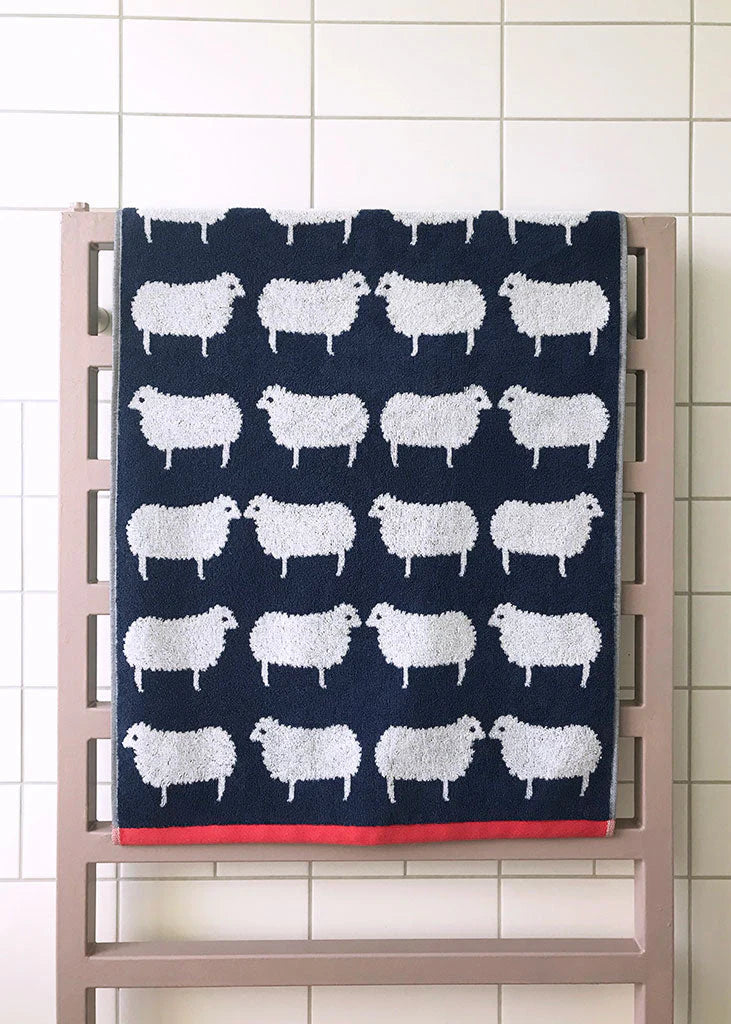 Sheep Organic Cotton Handtuch – dunkelblaues Baumwolltuch mit weißen Schafen, roter Saumkante, auf Heizkörper – Badetextilien
