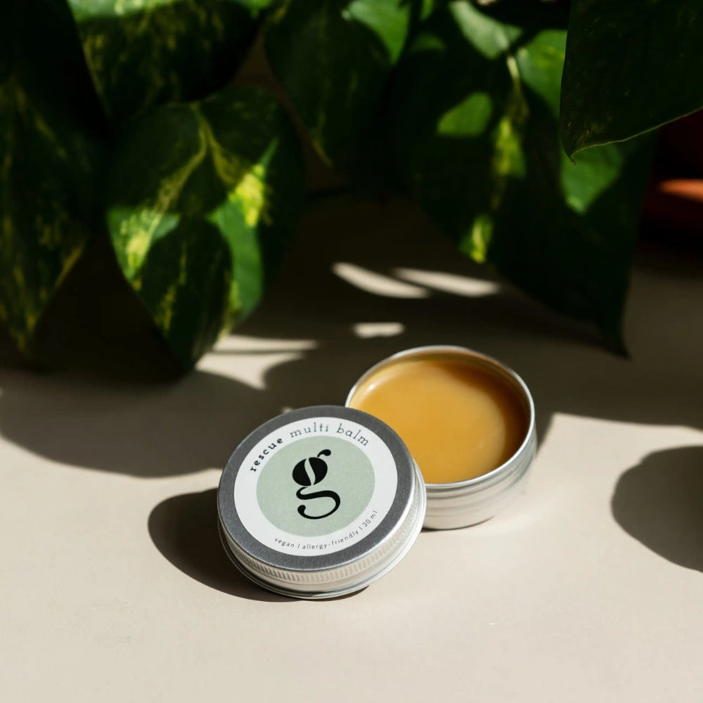 Rescue Multi Balm – silberner Metalldose 30 ml, geöffnet mit cremigem Balsam, beige Hintergrund mit grünen Blättern – Hautpflege