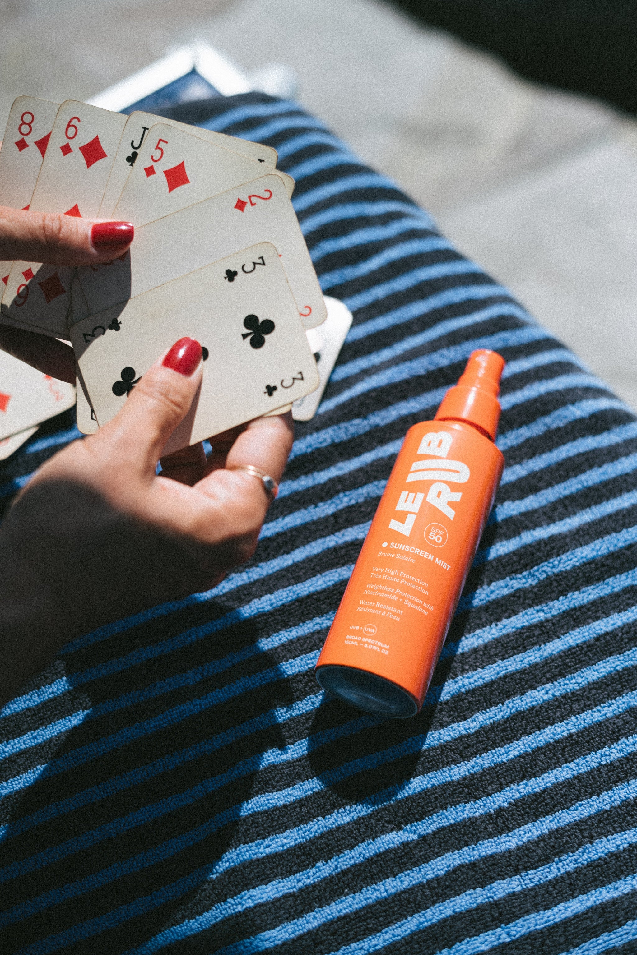 Orange LE RJ Sunscreen Mist SPF 50 spray bottle with cap on a blue and white towel, body sun care.