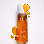RMS Beauty Kakadu Body Oil, goldfarbene Flasche mit weißem Verschluss und Textur vor weißem Hintergrund, Körperpflege