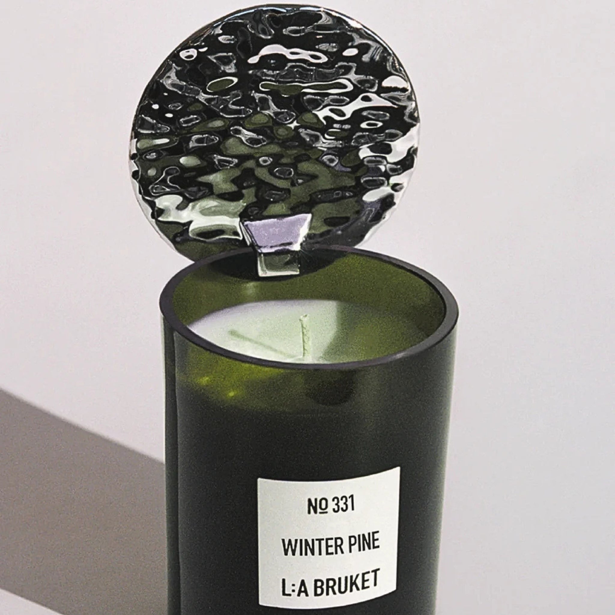 L:A Bruket Candle Reflektor, grünes Glasgefäß mit silbernem Metalldeckel vor hellem Hintergrund, Duftkerze.