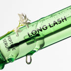 GreenGlam Long Lash Peptid-Wimpernserum: Nahaufnahme einer grünen Flasche mit Serum-Tropfen und kleinen weißen Blüten.