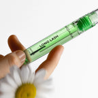 Mádara Long Lash Peptid-Wimpernserum in einer durchsichtigen grünen Tube, gehalten von einer Hand über einer Blume.