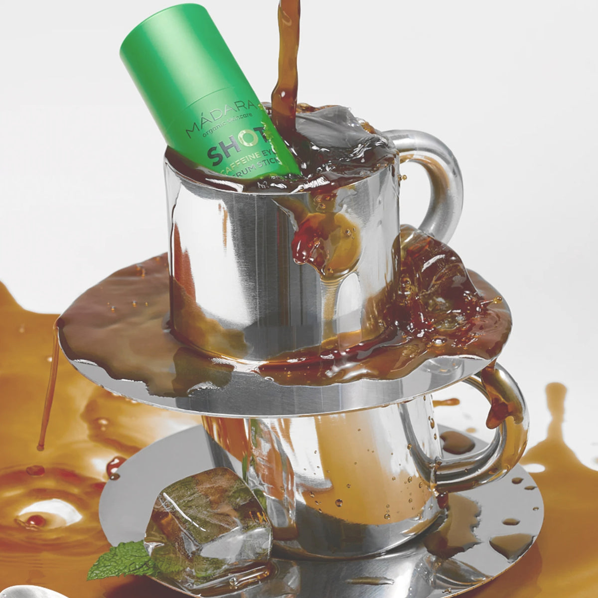 Mádara SHOT Koffein-Augenserum-Stick in einer grünen Stick, platziert in einer silbernen Tasse mit Kaffee-Spritzern.