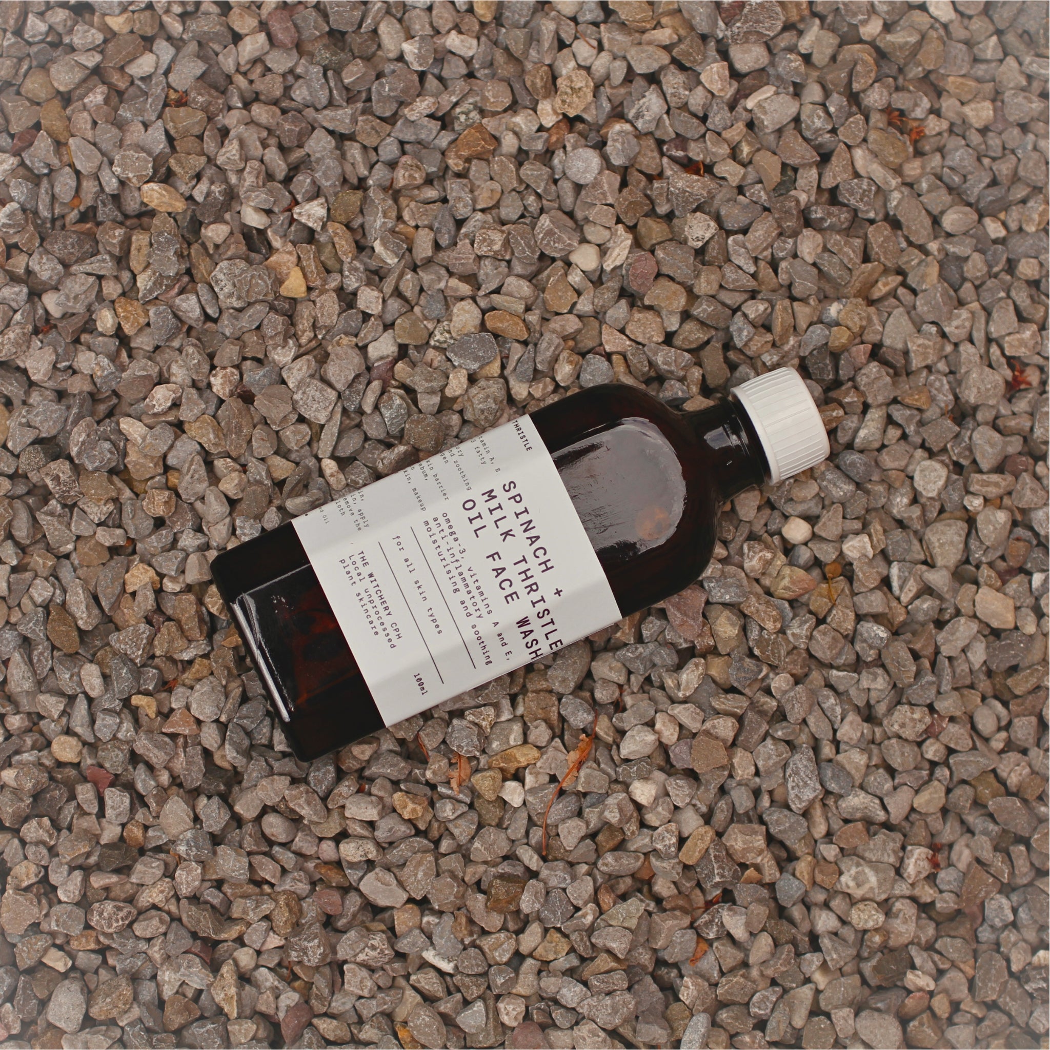 Spinach + Milk Thristle Oil Face Wash in einer braunen Glasflasche mit weißem Verschluss auf Kies, Hautpflege