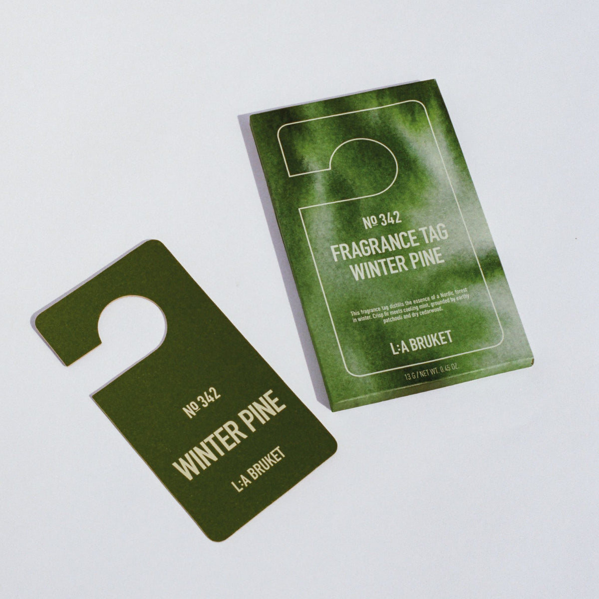 L:A Bruket No. 342 Fragrance Tag Winter Pine: ein dunkelgrüner Duftanhänger neben seiner Kartenverpackung.