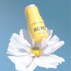 Mádara XOXO Lippenbalsam in einem gelben Stick, platziert auf einer weißen Blüte vor blauem Hintergrund.